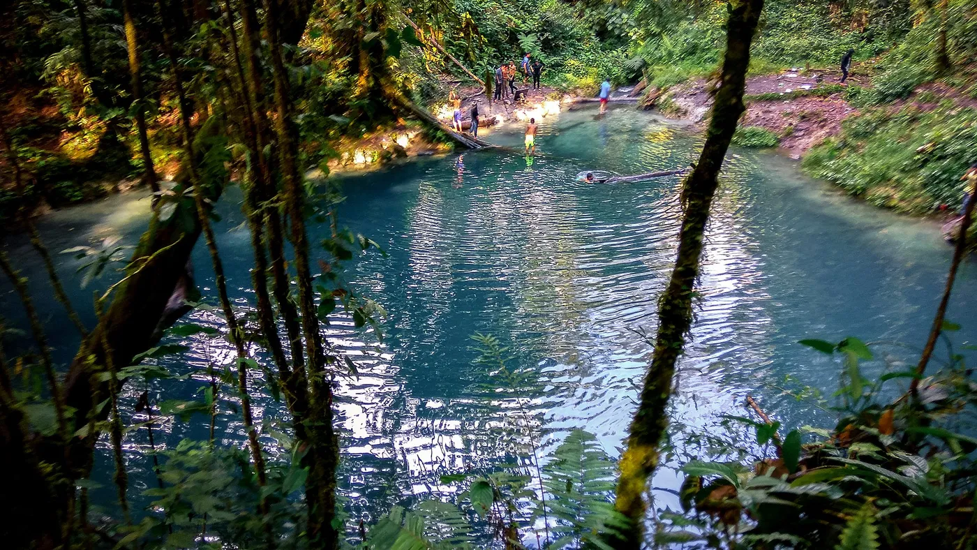 Blue water of Danau Kaco framed by tree trunks and rainforest in Kerinci, Jambi.