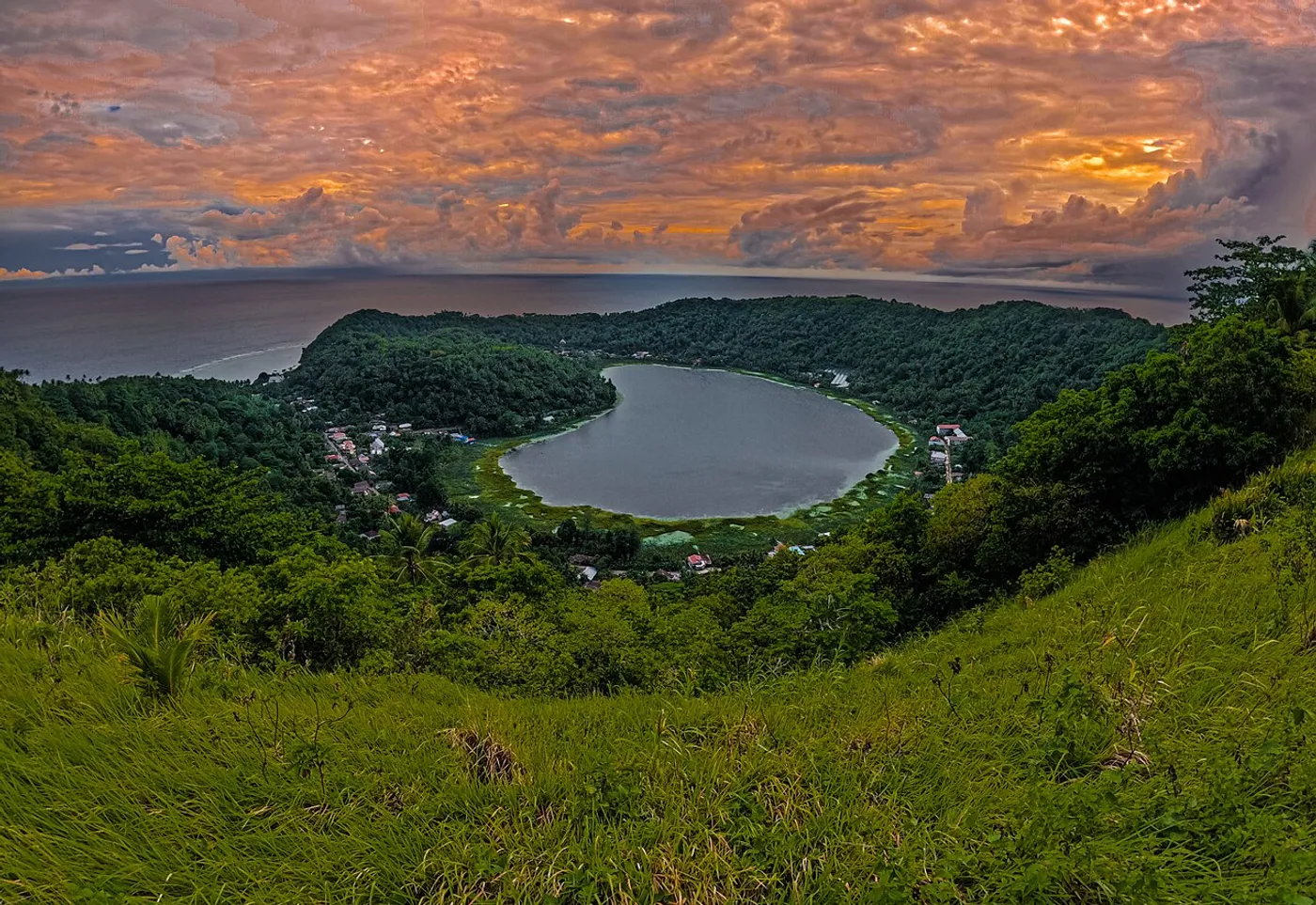 The heart-shaped Danau Cinta surrounded by green hills on Makalehi Island, with the sea and a colourful sky beyond