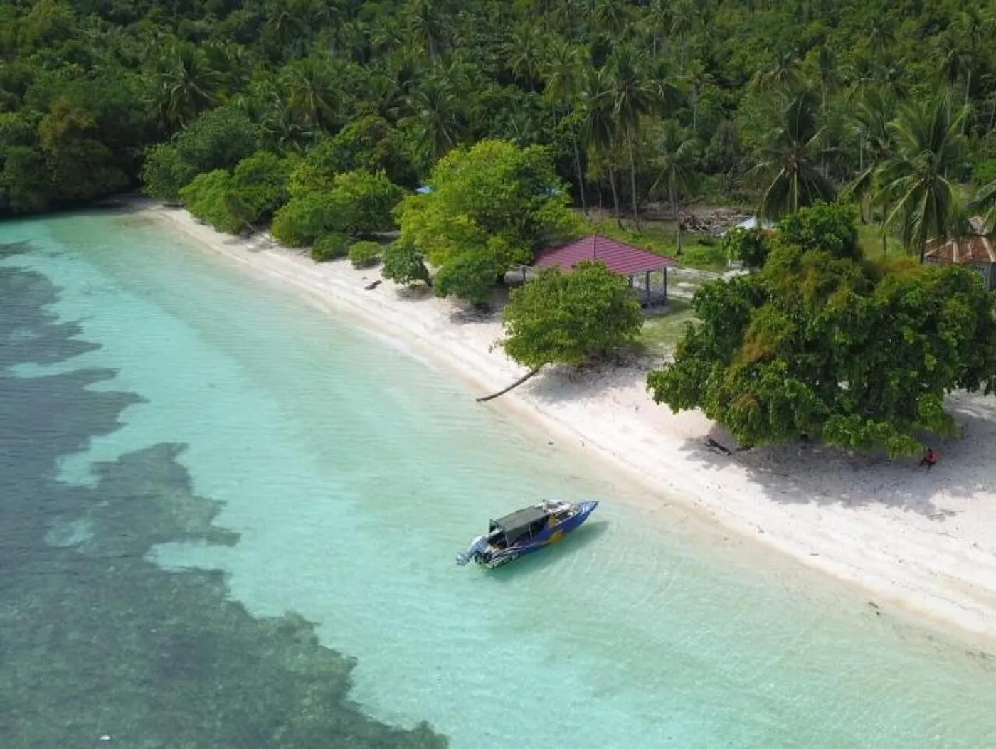 A small boat floats beside the white sand and palm-fringed shore of Pasir Panjang Beach on Mbuang-Mbuang Island